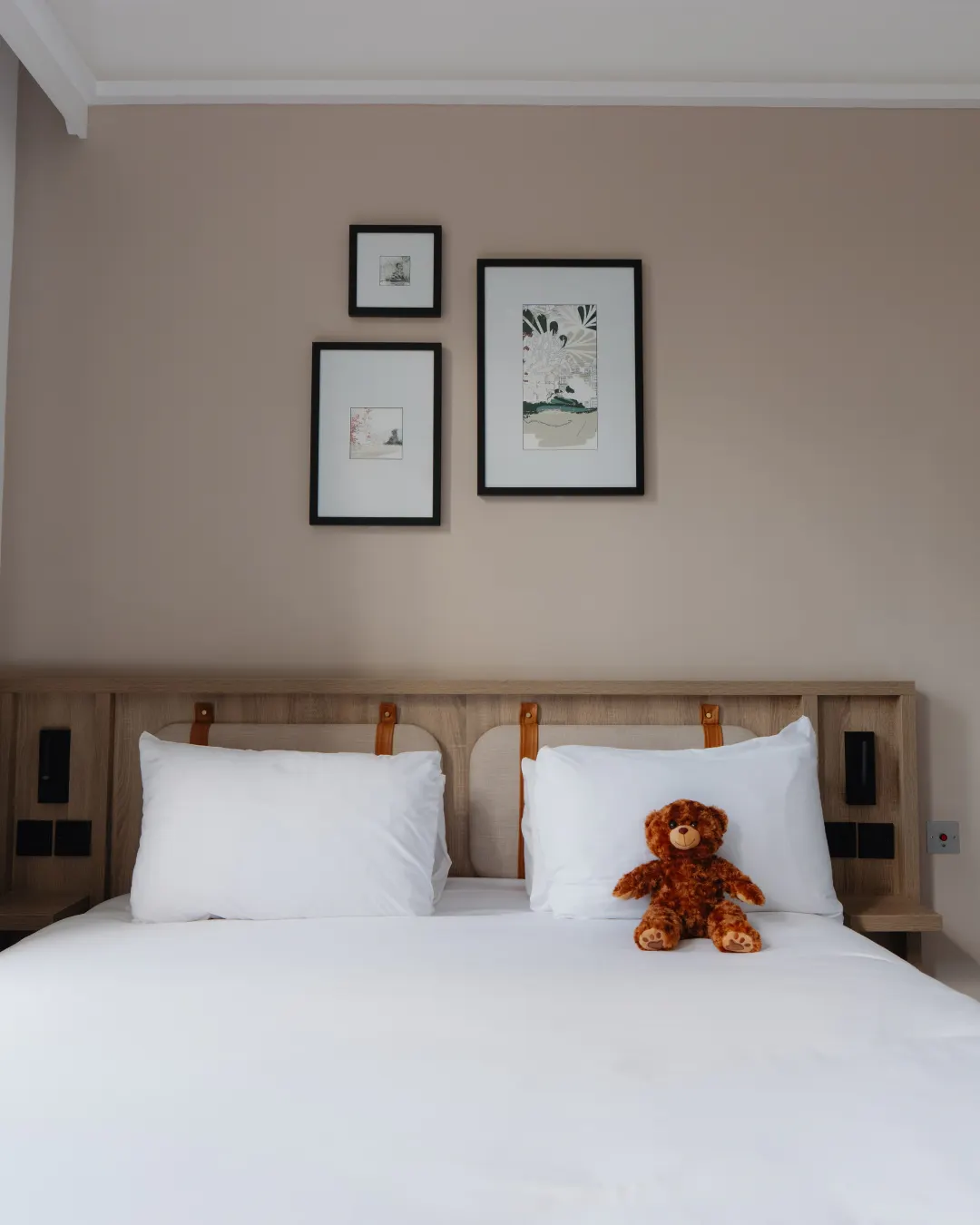 A teddy bear sits on a white hotel bed below three framed artworks.