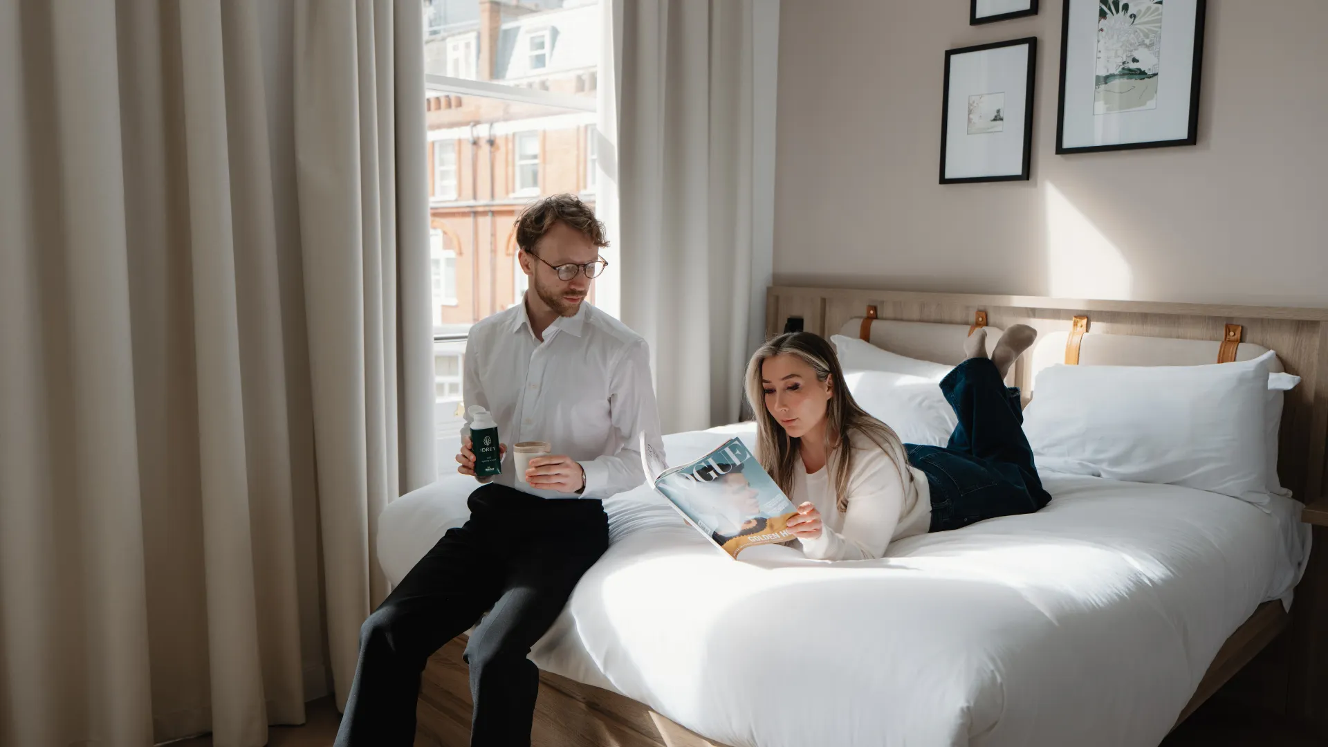 Two guests relax on a white bed in a bright hotel room with tall curtains and framed artwork on the wall.