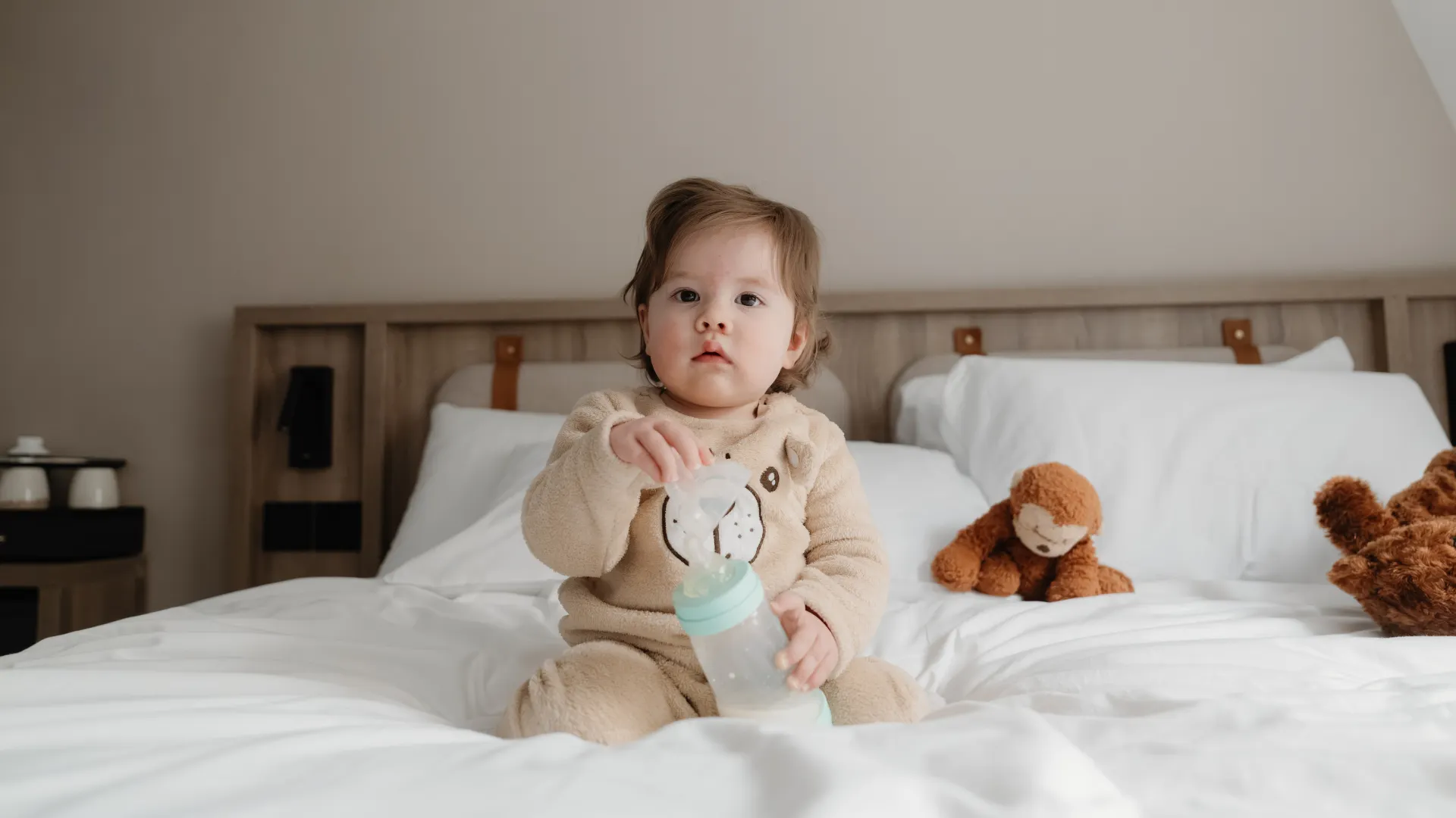 A toddler sits on a white hotel bed holding a baby bottle with stuffed toys beside them.