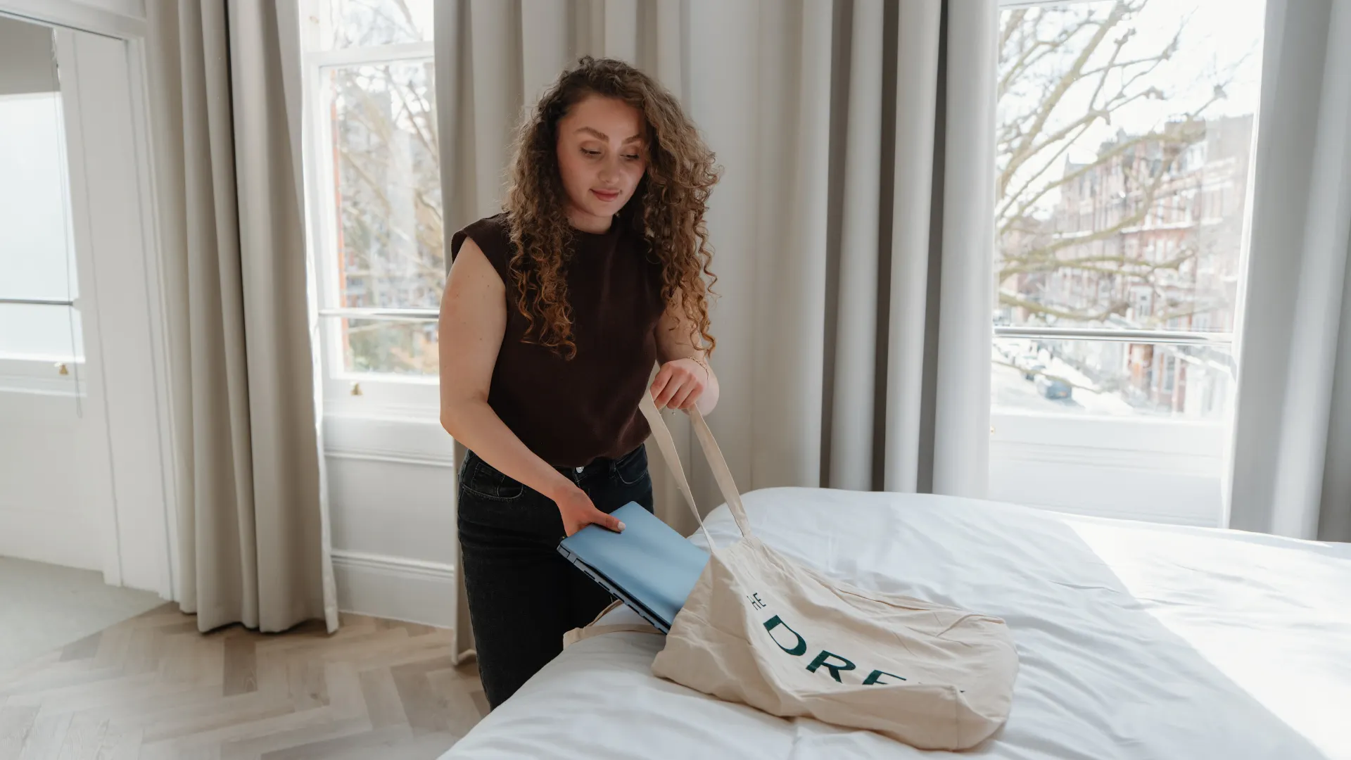 A guest unpacks a blue laptop from a canvas tote on a white bed beside tall windows. Visible text reads "DREY".