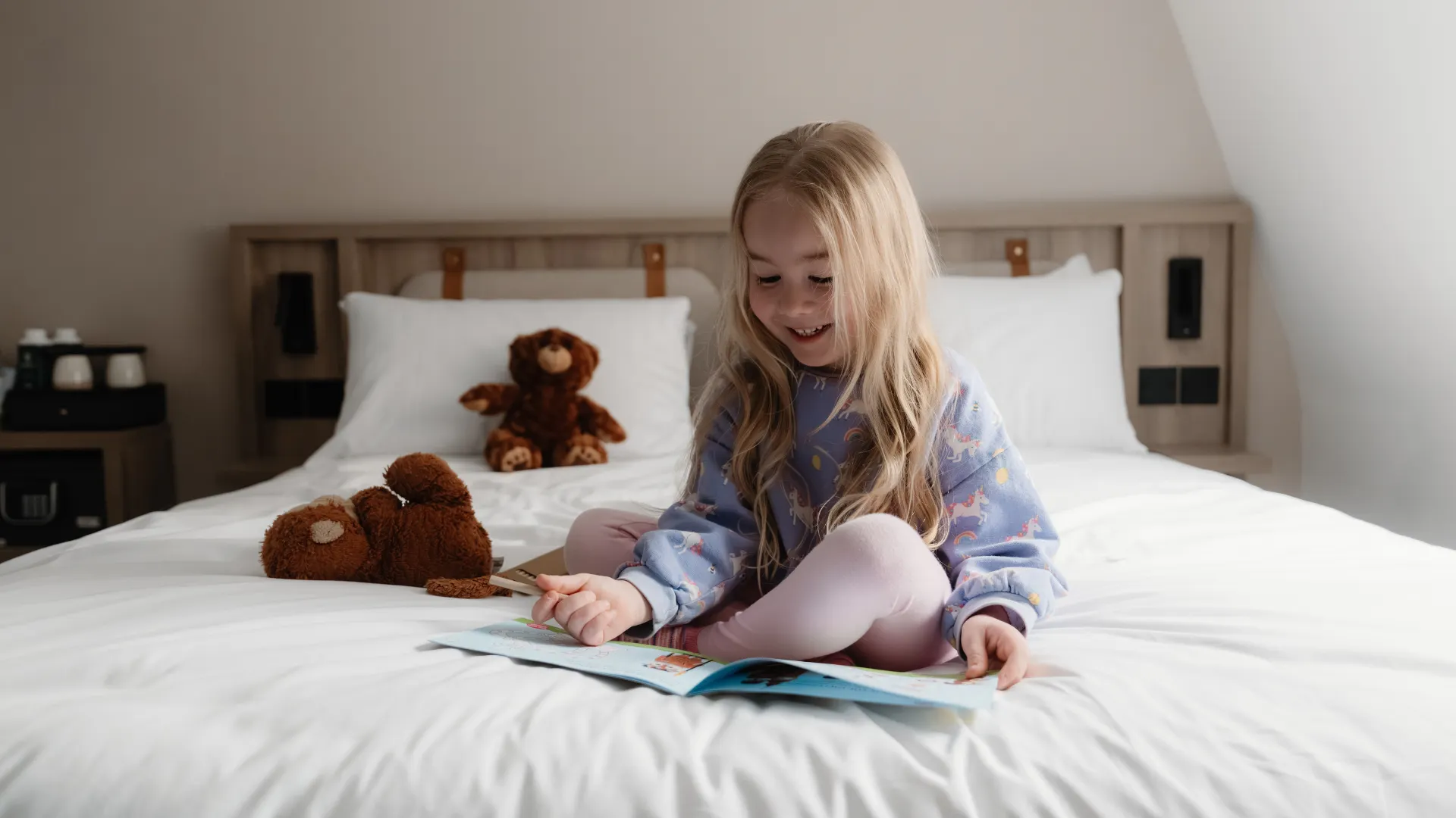 A smiling young girl reads a book on a white hotel bed with teddy bears beside her.