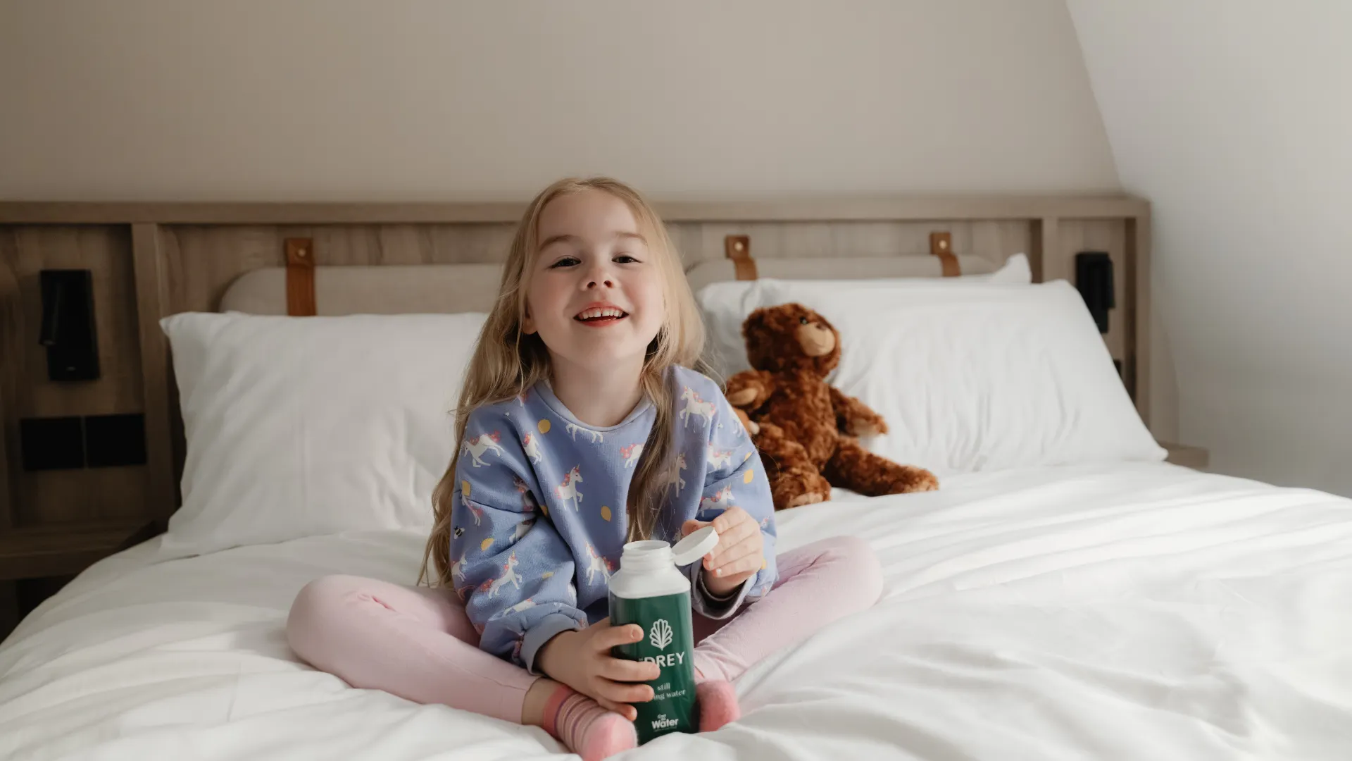 A young girl sits on a white hotel bed holding a carton of water with a teddy bear behind her. Visible text reads "THE DREY" and "still spring water".