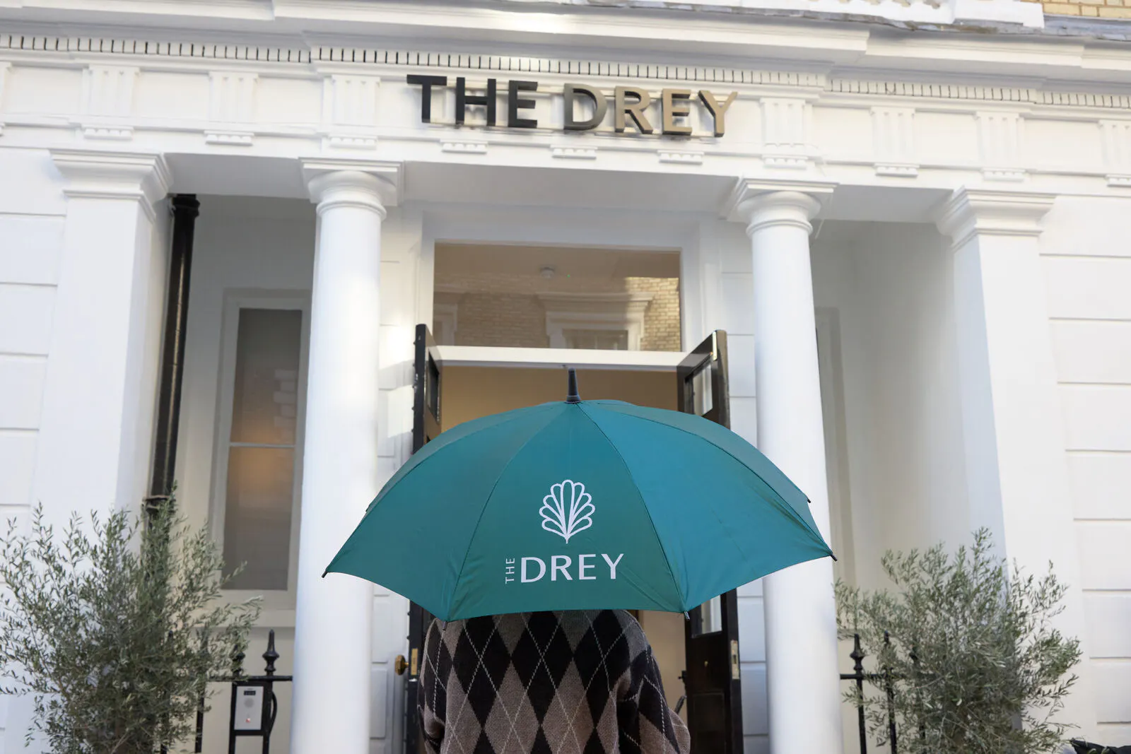 A guest stands under a teal umbrella outside The Drey entrance. Visible text reads "THE DREY".