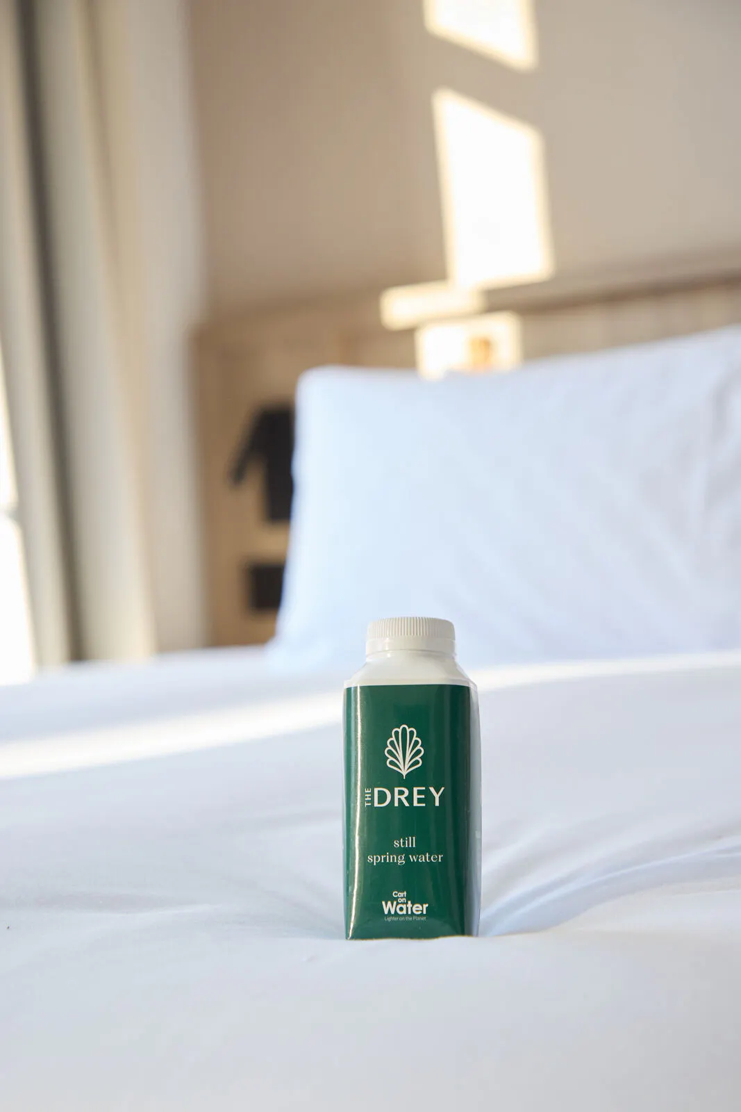 A carton of spring water stands on a white hotel bed in soft sunlight. Visible text reads "THE DREY" and "still spring water".