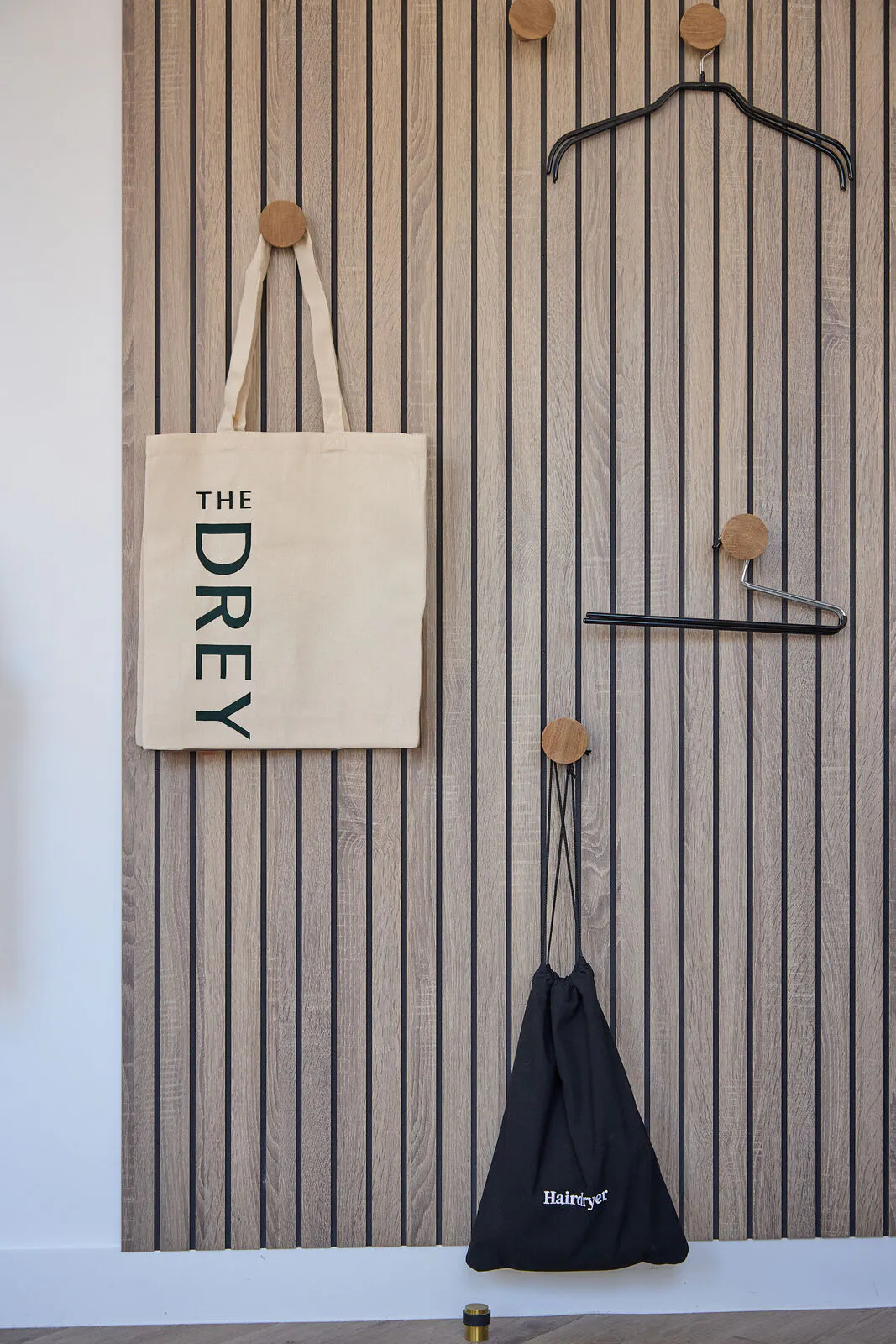 A canvas tote and black hairdryer bag hang on wooden wall hooks beside empty hangers. Visible text reads "THE DREY" and "Hairdryer".