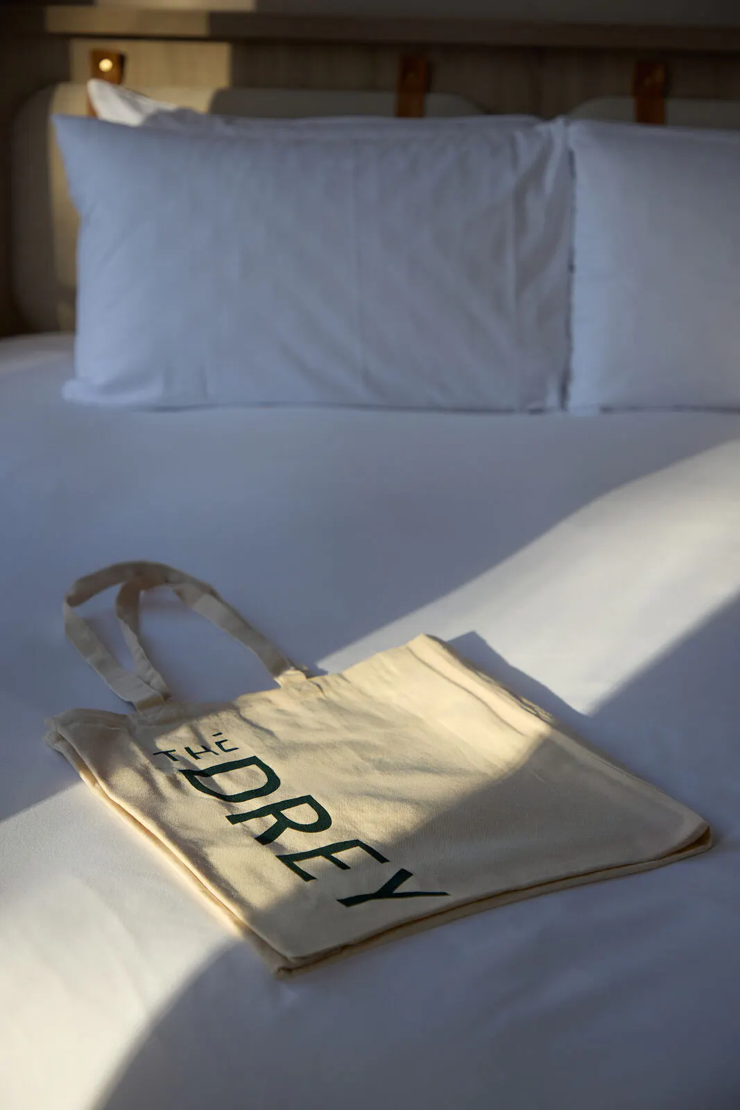 A folded canvas tote bag lies in sunlight on a white hotel bed. Visible text reads "THE DREY".
