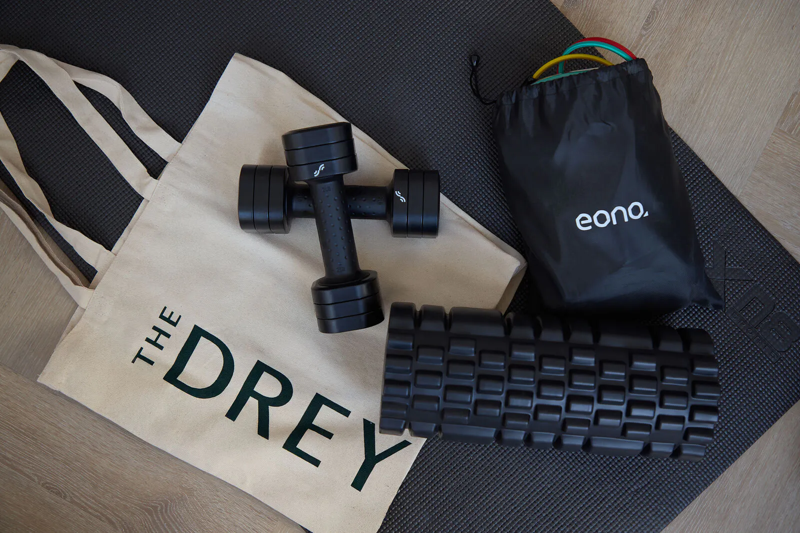 The Drey in room workout kit laid out on a black exercise mat with dumbbells a foam roller resistance bands and a tote bag. Visible text reads "THE DREY" and "eono."