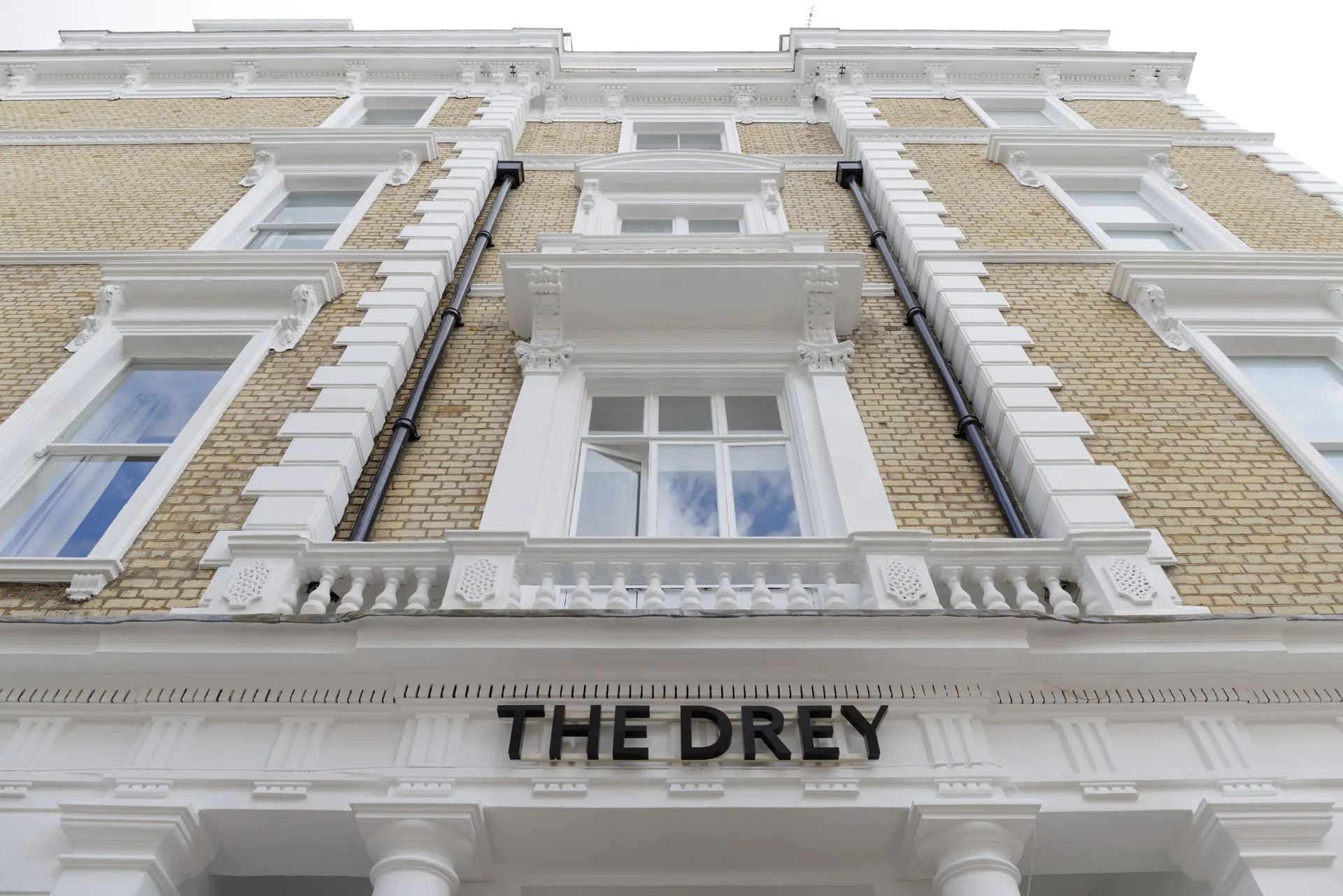 Low angle view of The Drey exterior showing cream brickwork white trim and the hotel sign above the entrance. Visible text reads "THE DREY".