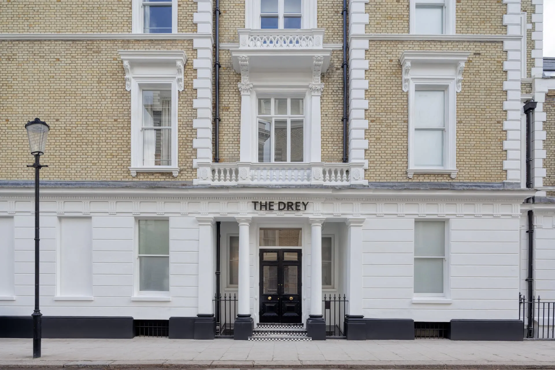 Straight on view of The Drey exterior with cream brickwork white columns black railings and a quiet pavement. Visible text reads "THE DREY".