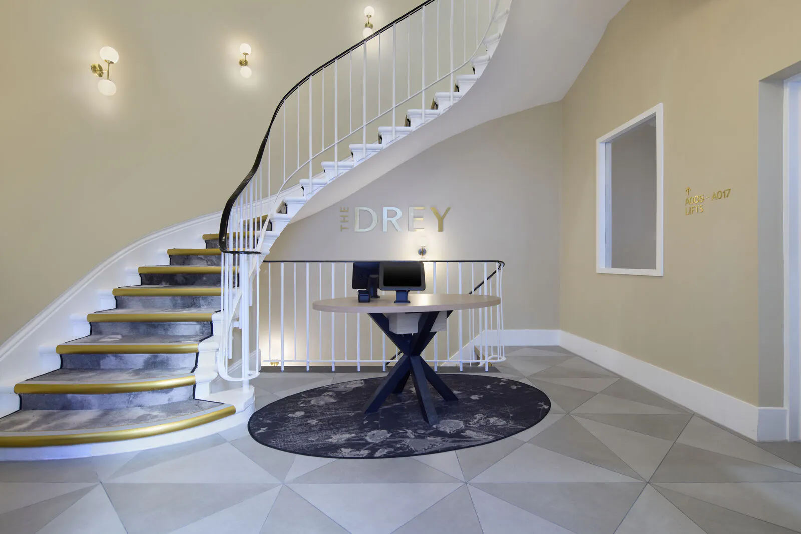 The Drey reception area with a round check in table beneath a curved staircase. Visible text reads "THE DREY" and "A105 A017 LIFTS".