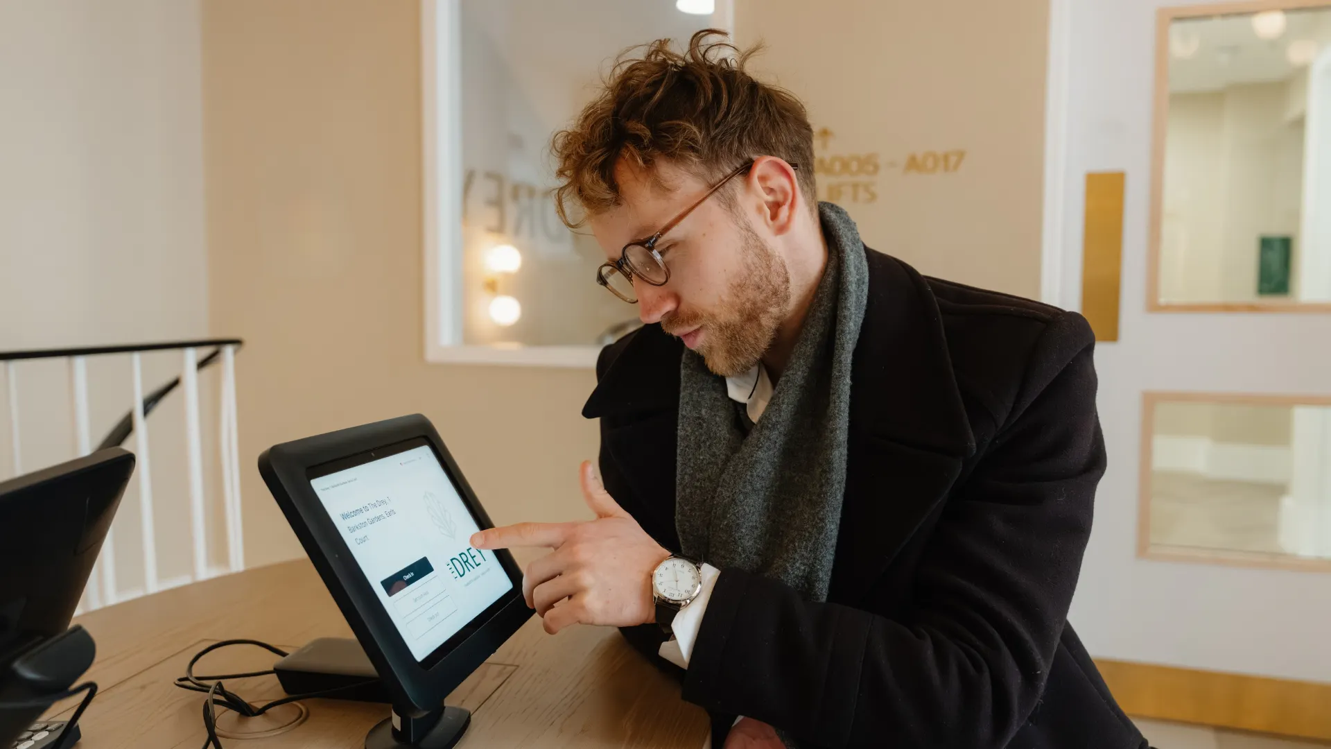 A guest uses a tablet at The Drey check in desk with room numbers visible in the background. Visible text reads "A005 A017 LIFTS" and "DREY".