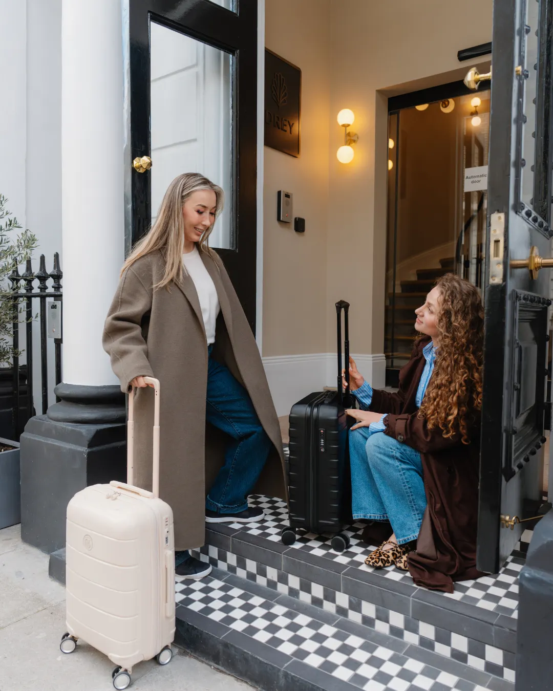 Two guests with suitcases pause on The Drey entrance steps beside open black doors.