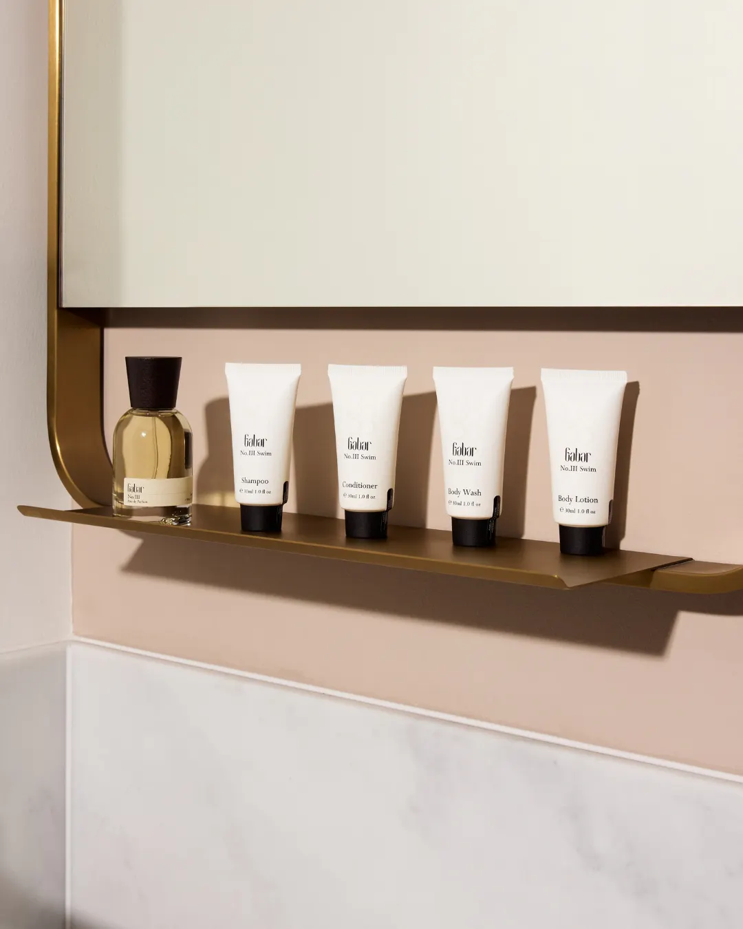 Gabar fragrance and toiletry products sit on a brass bathroom shelf beneath a mirror. Visible text reads "Gabar No.III Swim Shampoo Conditioner Body Wash Body Lotion".