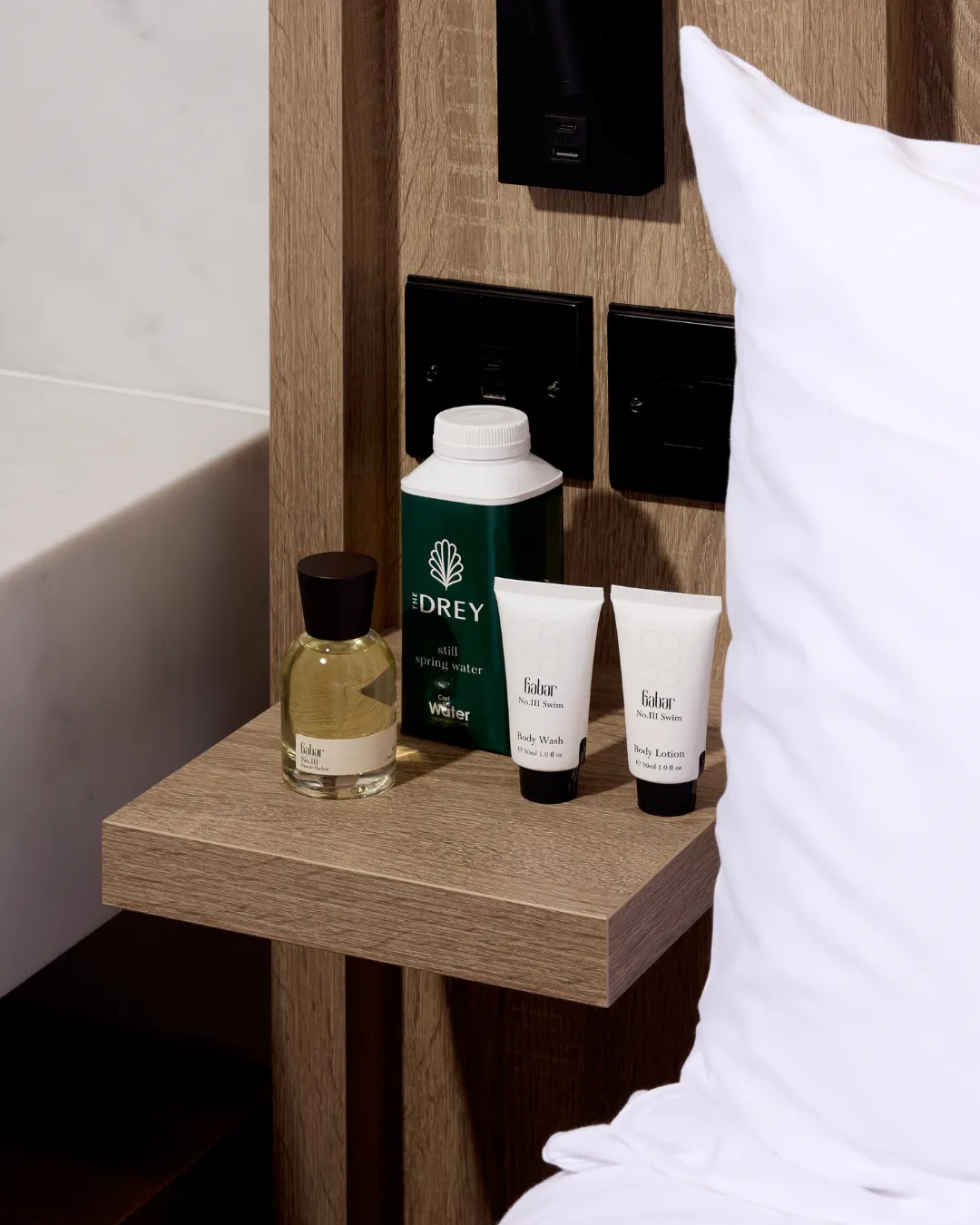A bedside shelf holds Gabar products and a carton of spring water beside a white pillow. Visible text reads "THE DREY" and "Gabar No.III Swim Body Wash Body Lotion".