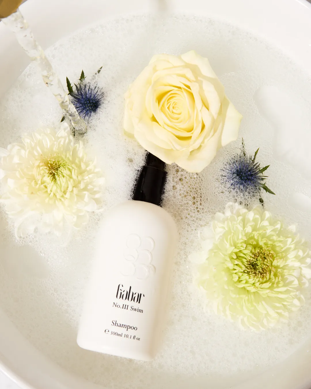 A Gabar shampoo bottle rests in bath bubbles with pale flowers and blue thistles around it. Visible text reads "Gabar No.III Swim Shampoo".
