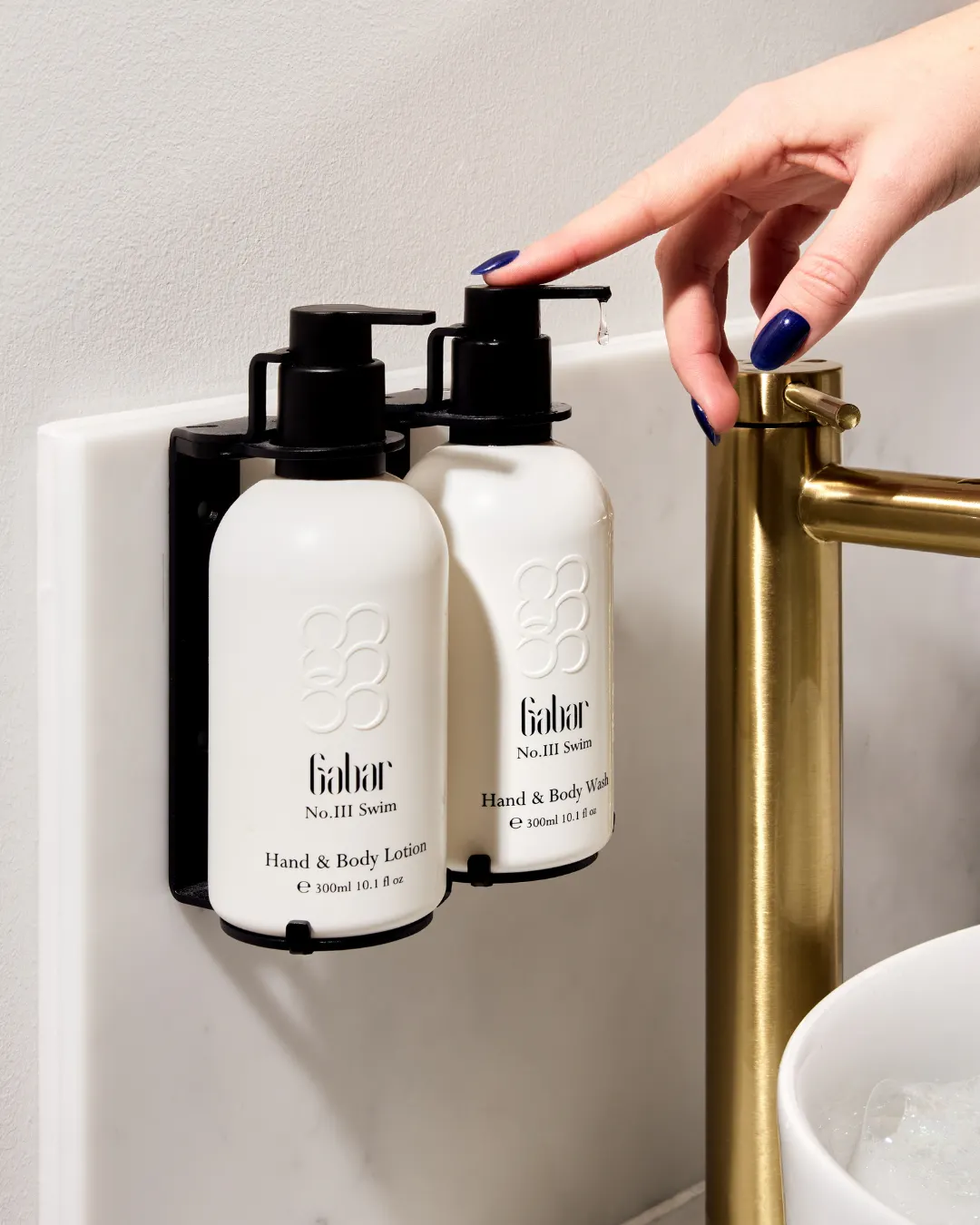 A hand presses the pump on a refillable Gabar bottle beside a brushed gold bathroom tap. Visible text reads "Gabar No.III Swim Hand and Body Lotion" and "Gabar No.III Swim Hand and Body Wash".