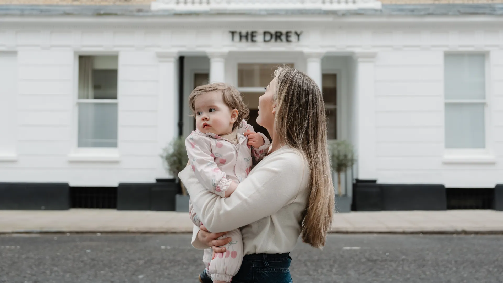 A woman holding a young child stands in front of a white building with the sign "THE DREY" above the entrance.