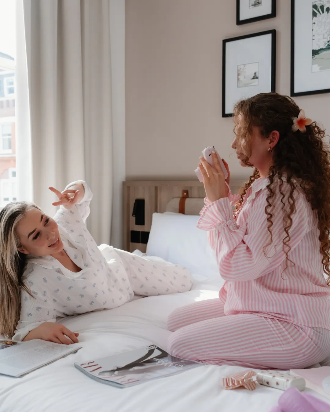 Two guests in pyjamas on a hotel bed, one taking a Polaroid photo while the other poses, with a magazine on the bed.