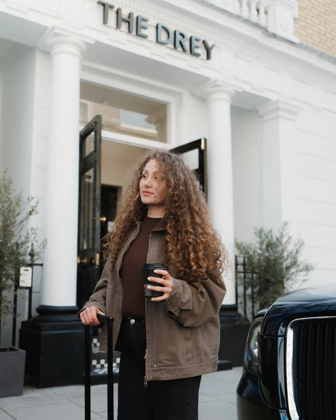 A young woman stands outside The Drey entrance in front of a black taxi, holding a coffee while resting her hand on her black suitcase.