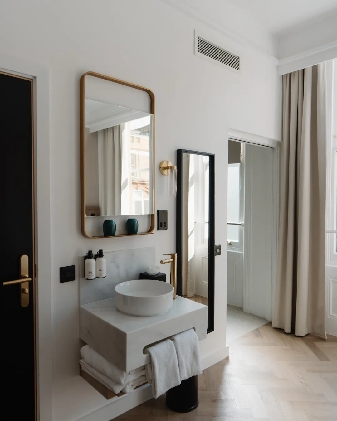 Modern bathroom with a floating marble sink, round vessel basin, large rectangular mirror, toiletries, towels, and floor-to-ceiling curtains over tall windows.