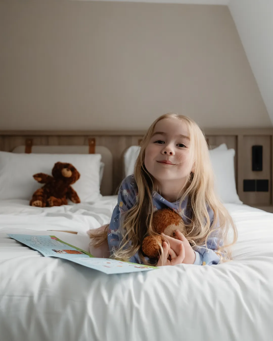 A young girl with long blonde hair lies on a bed, holding a stuffed animal and a book, with another teddy bear placed on a pillow in the background.