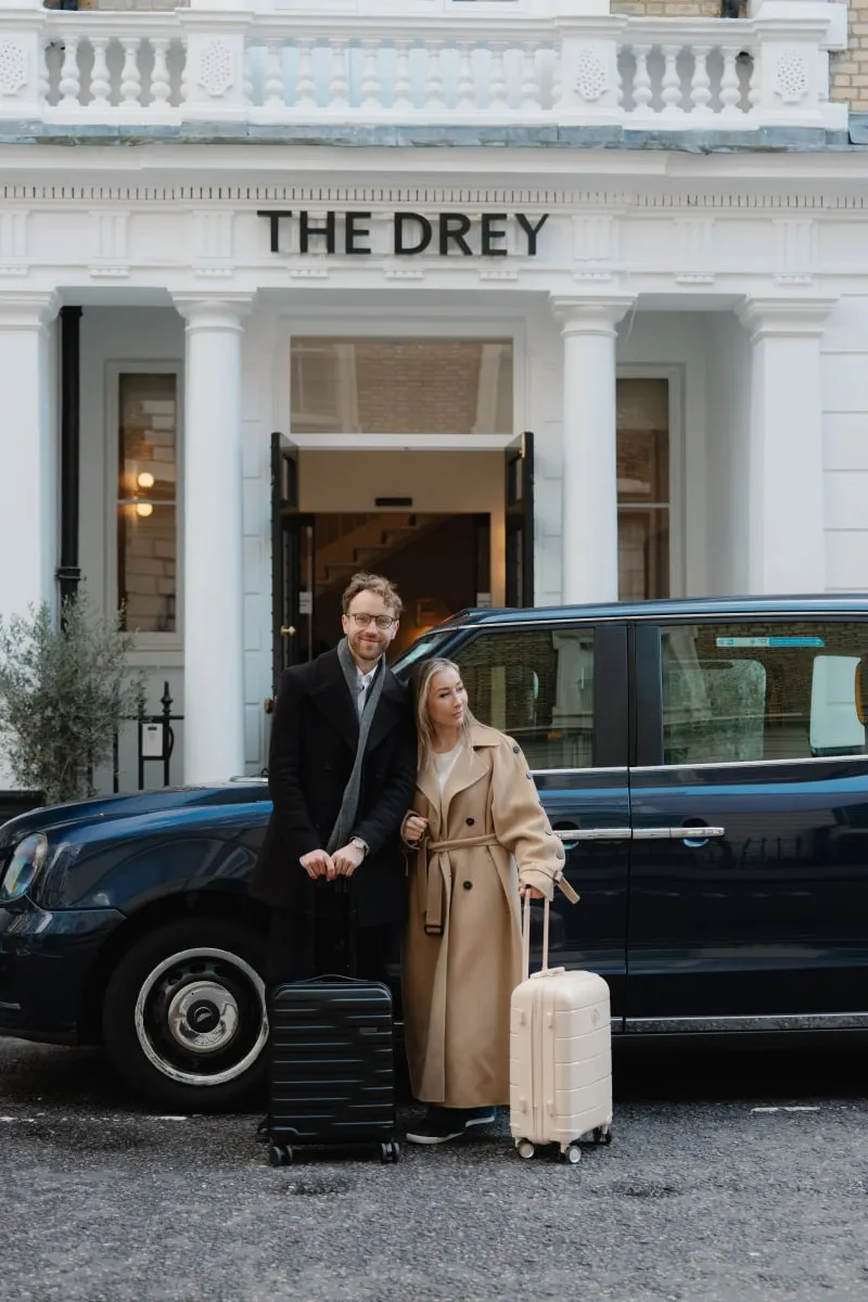 Two guests stand with suitcases beside a black taxi outside The Drey entrance. Visible text reads "THE DREY".