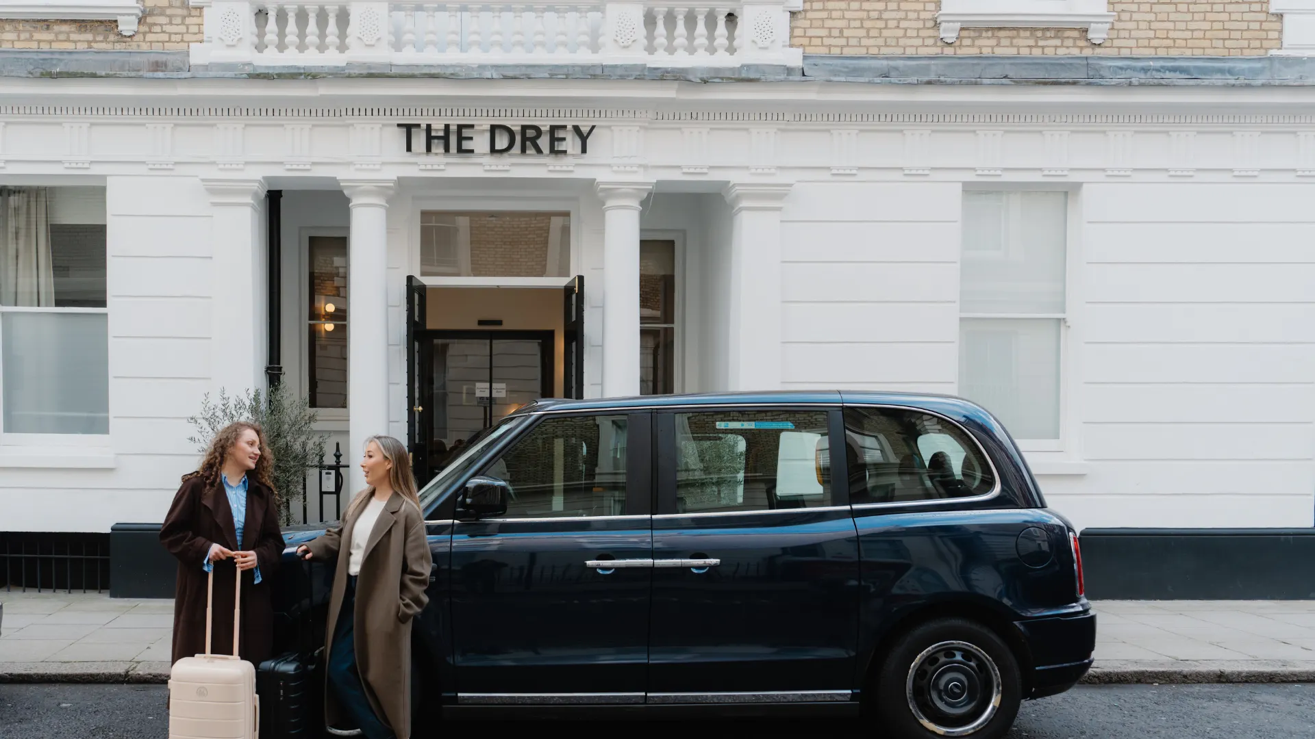 Two women with luggage stand beside a black taxi in front of a white building with a sign reading "The Drey".